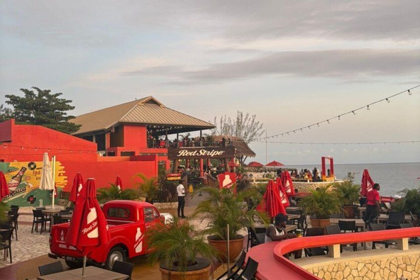 A beachside restaurant with outdoor seating, a red truck, and a view of the sea. 1