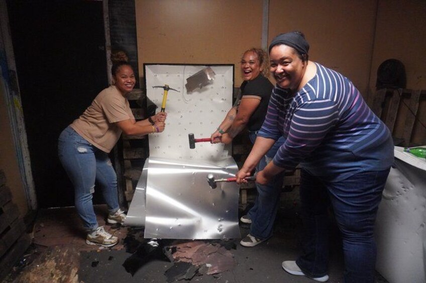 Four people working on a large metal object, possibly a door or panel, in an indoor setting with tools and debris on the floor. 4