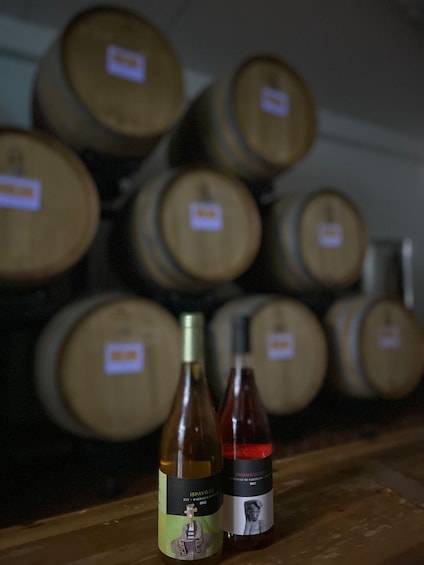 Two wine bottles in front of a row of wooden barrels. 2