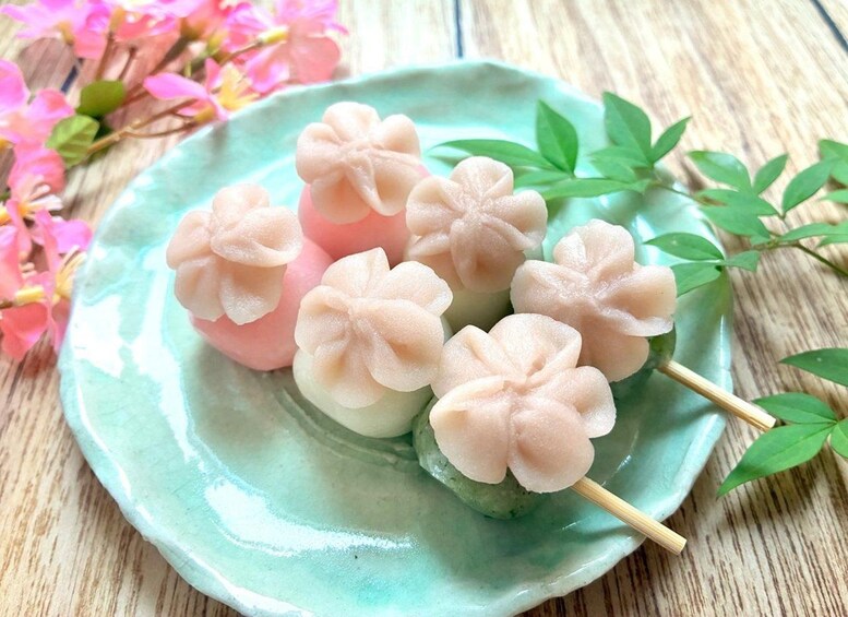 A plate of pink and white mochi with flower-shaped toppings, placed on a green ceramic plate with a wooden background. 2