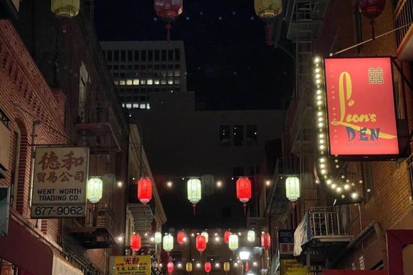 A street at night with illuminated signs, including a prominent red sign for 'Lion's Den Bar Lounge,' and traditional red lanterns hanging overhead. 3