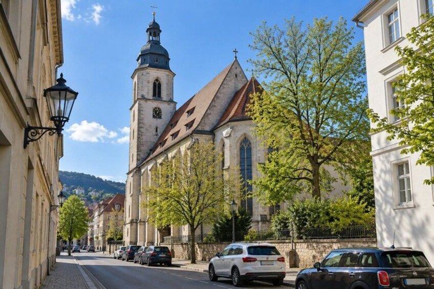 A street with parked cars, a church with a tall tower, and trees lining the road. 5