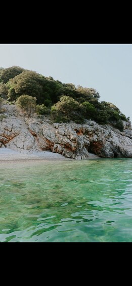 A rocky shoreline with clear turquoise water and dense green foliage on a hill. 2
