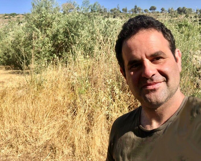 A person standing in a field with dry grass and bushes in the background. 2