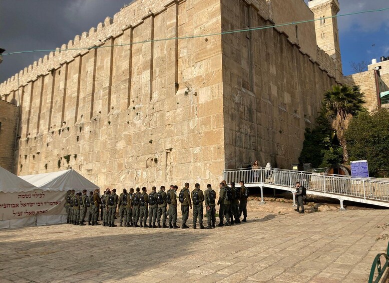 A group of soldiers standing in formation in front of a large stone wall with a guard tower. 1