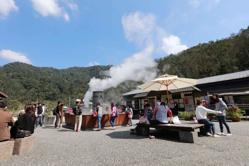 A group of people gathered around a steam vent, with a traditional building in the background. 3