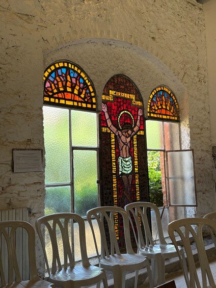 Interior de iglesia con vidrieras que representan figuras religiosas, sillas de plástico blanco y una pared de piedra. 5