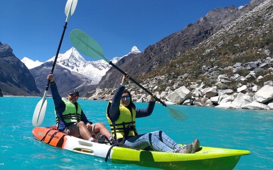 Two people kayaking on a turquoise lake with snow-capped mountains in the background. 1