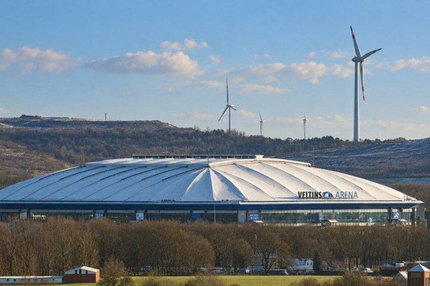 A large sports stadium with a curved roof, surrounded by trees and hills, with wind turbines in the background. 3