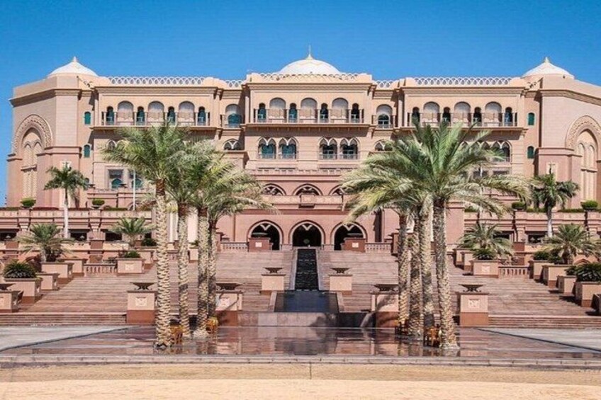 A large, multi-story building with a central entrance, palm trees, and a clear blue sky. 3