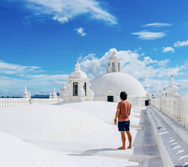 A person standing on a white rooftop overlooking a white dome structure with multiple smaller domes and spires, under a clear blue sky. 1