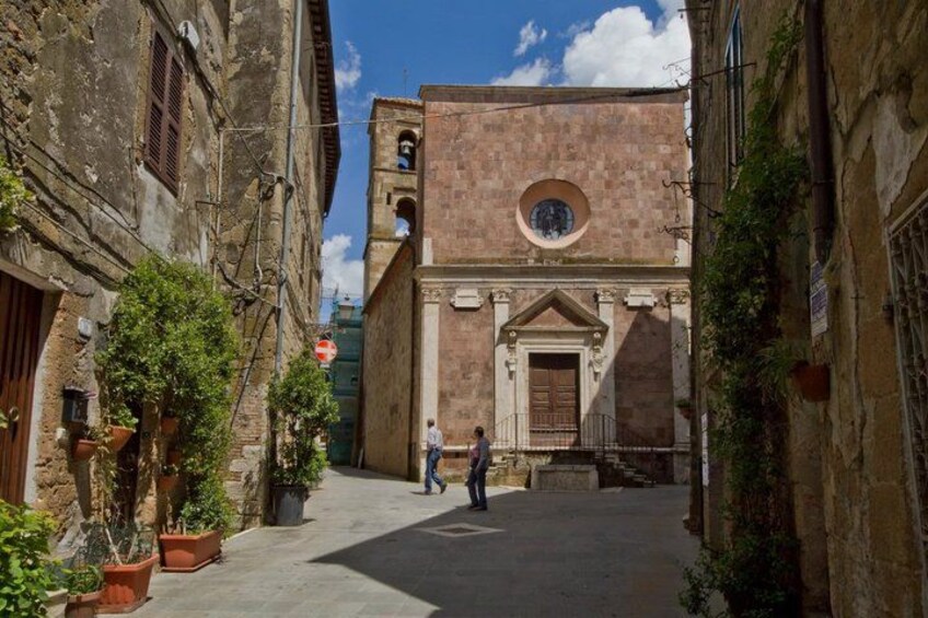 A narrow street with historic buildings, a bell tower, and potted plants. 4