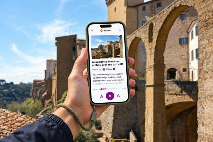 A hand holding a smartphone displaying a photo of Acquedotto Mediceo arches over the tuff cliff with a historic building in the background. 1