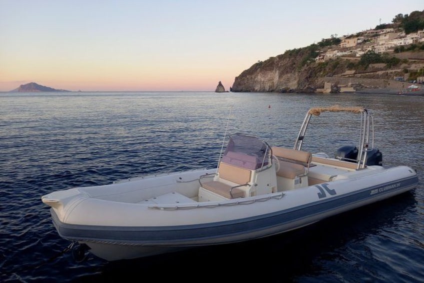 A small motorboat on calm water with a rocky coastline and buildings in the background. 1