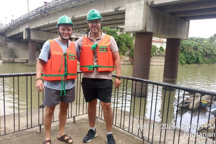 Two people wearing life vests and helmets standing by a river with a bridge in the background. 2