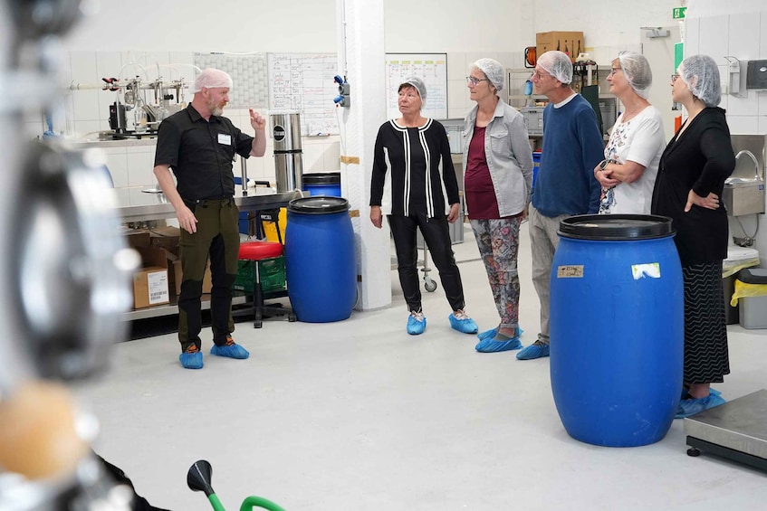 A group of people in a laboratory setting with large blue barrels, white walls, and various scientific equipment. 4