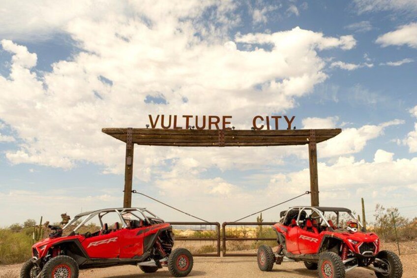 Two red off-road vehicles parked under a wooden archway with the text 'VULTURE CITY' on it. 5