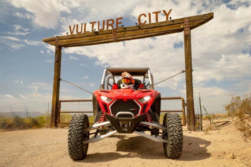 A red off-road vehicle at a wooden archway with the words 'VULTURE CITY' on it. 1