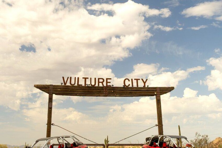 A wooden sign with 'VULTURE CITY' written on it, two cars parked below, and a clear sky with clouds. 3