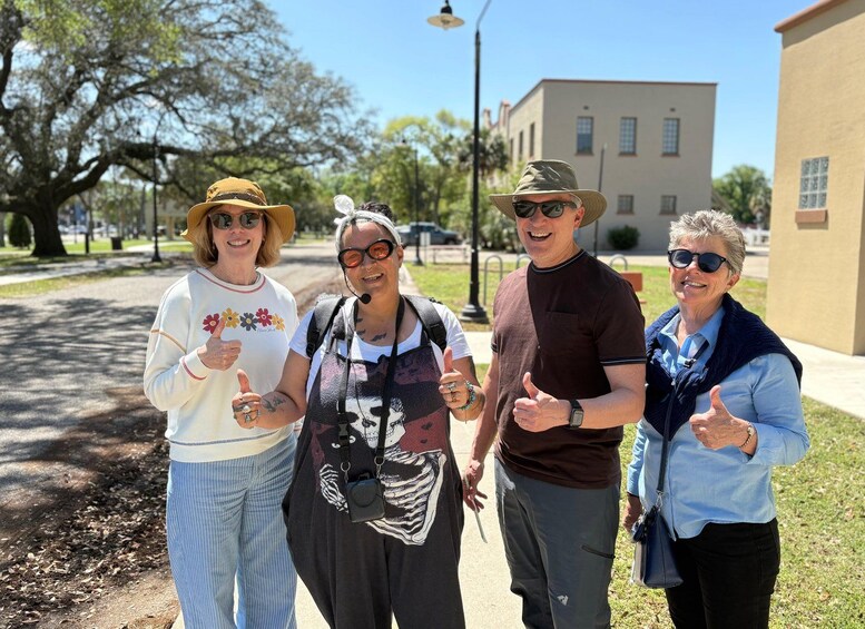 Four people giving thumbs up in a sunny outdoor setting with trees and buildings in the background. 1