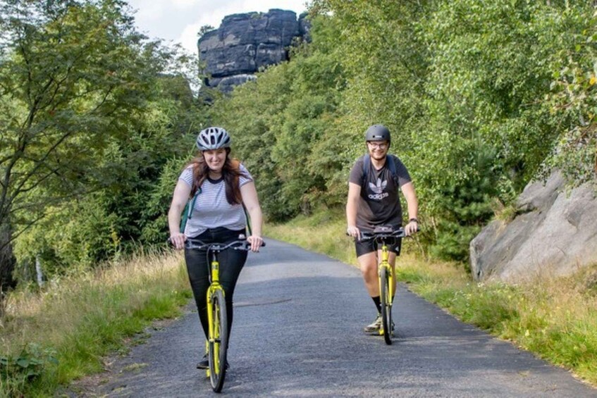 Two people riding bicycles on a paved path surrounded by greenery and a large rock formation in the background. 2