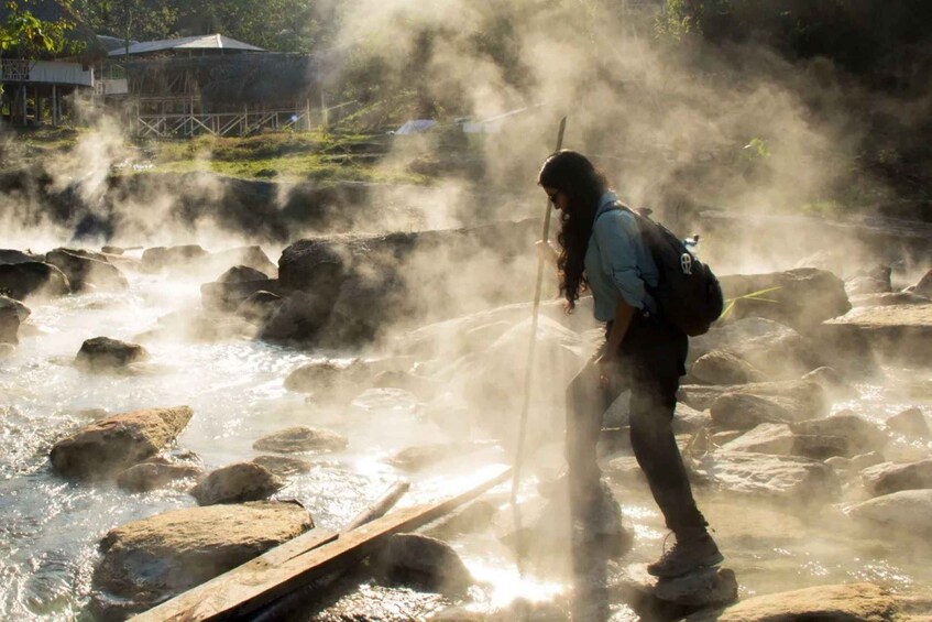 Une personne traverse une rivière où s’élève de la vapeur, entourée de rochers et avec un pont en arrière-plan. 4