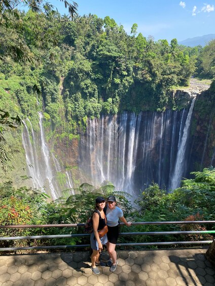 A waterfall with lush greenery and a paved viewing area. 2