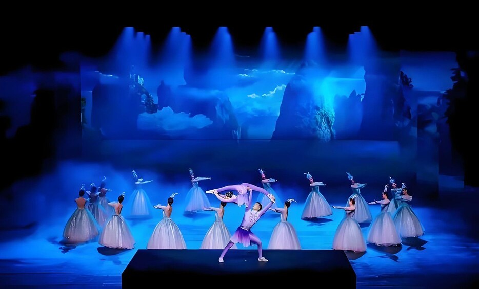 Un spectacle de ballet avec des danseurs vêtus de costumes blancs et violets, sur une scène dont le fond est illuminé en bleu. 1