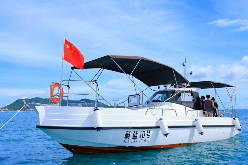Ein weißes Boot mit einer roten Flagge und chinesischen Schriftzeichen an der Seite, das auf blauem Wasser unter einem wolkenlosen Himmel fährt. 1