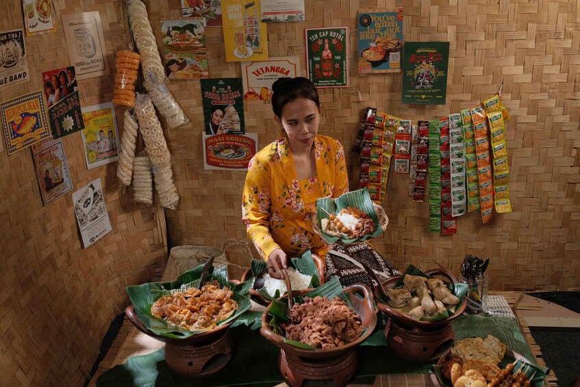 A woman serving food at a stall with various food items and promotional posters on the wall. 1