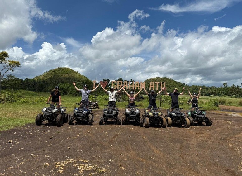 Fünf Personen auf Quads mit erhobenen Armen, im Hintergrund ein Schild mit der Aufschrift 'ICRAHAM ATV' und ein bewölkter Himmel. 3