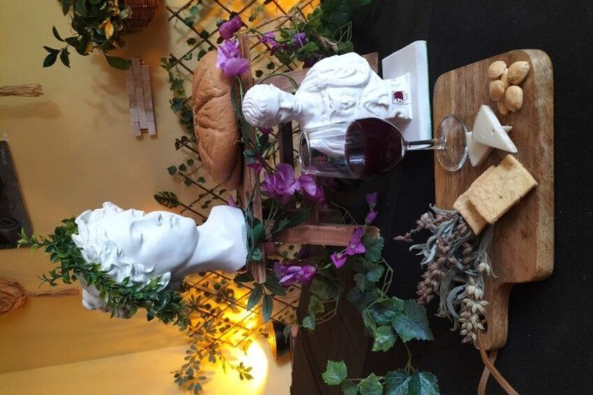 A table with a bust sculpture, a glass of red wine, cheese, and a cutting board with nuts. 3
