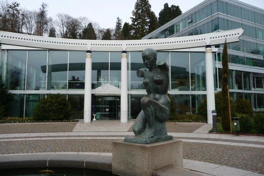 A modern building with large glass windows, a central sculpture, and a paved area in front. 3