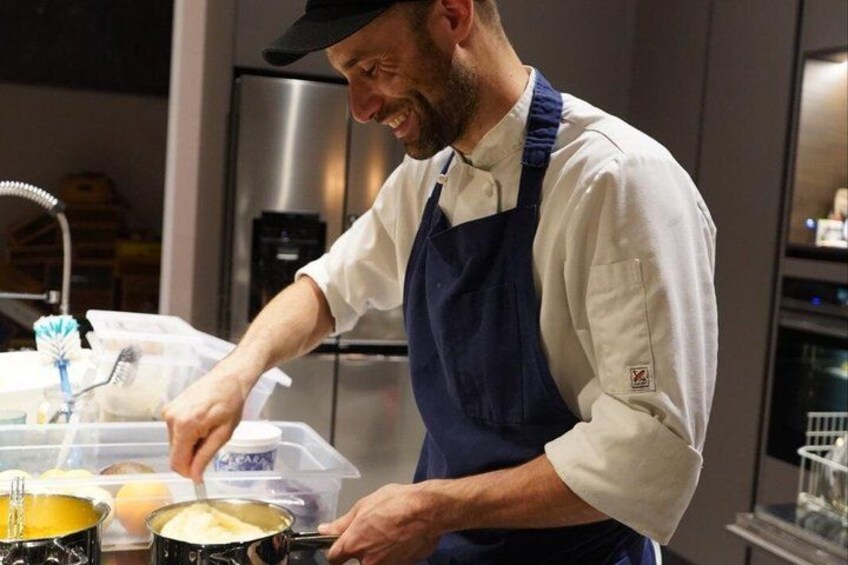 A person in a white shirt and blue apron cooking in a kitchen with pots and a refrigerator in the background. 1