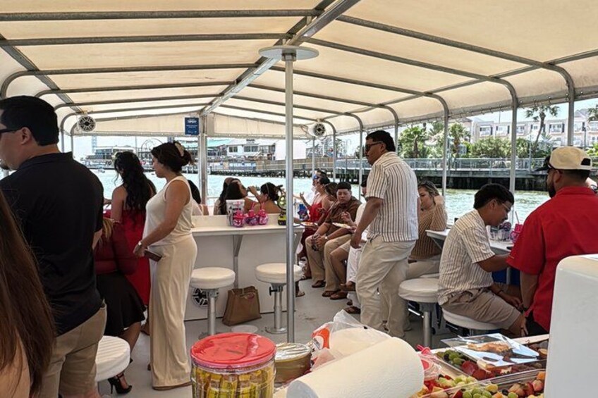 A covered boat with people sitting and standing, a bar area, and a view of the water and buildings in the background. 3
