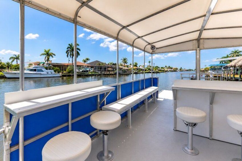 A covered outdoor seating area with white bar stools and a blue railing overlooking a marina with boats and palm trees. 5