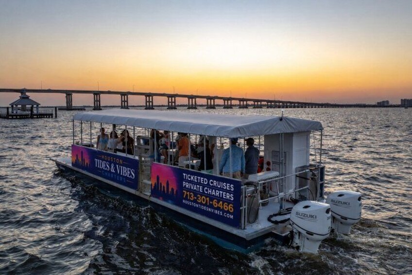 A boat with 'Houston Tides & Vibes' and 'Ticketed Cruises Private Charters' on its side, docked at sunset with a pier in the background. 1