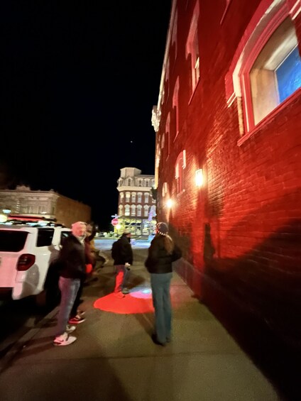 A nighttime street scene with a red brick building, a white van, and people walking on a sidewalk. 3