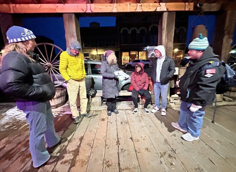 A group of people standing on a wooden deck at night, with a building and parked cars in the background. 5