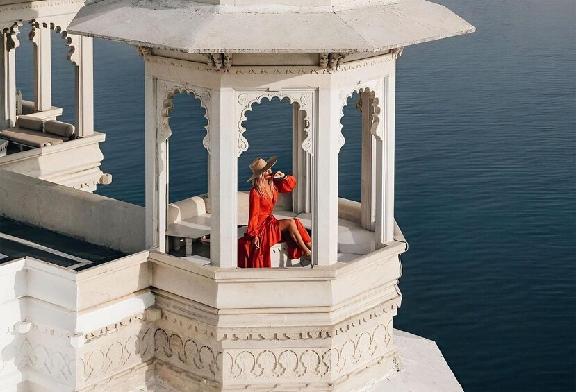A woman in a red dress sitting on a white bench under a gazebo with ornate white pillars, overlooking a body of water. 1