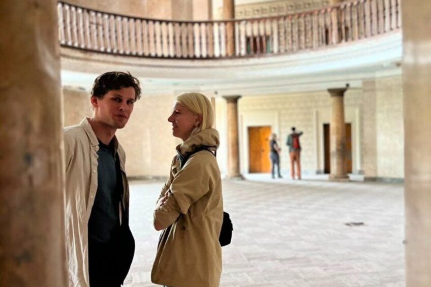 A man and a woman in a conversation inside a building with a high ceiling and columns. 4