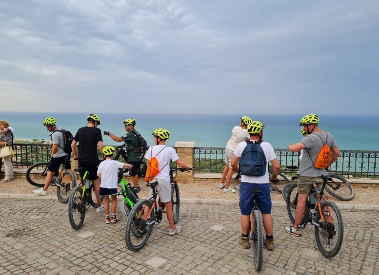 A group of people on bicycles with helmets, standing on a paved area with a railing, overlooking a body of water. 1