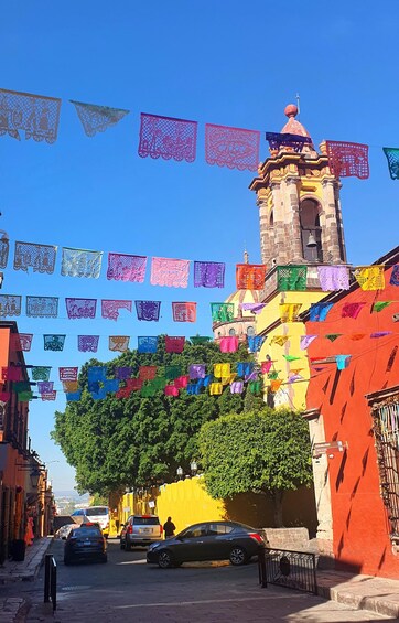 A street with colorful papel picado flags, a church with a bell tower, and parked cars. 2