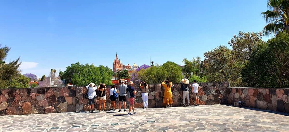 A group of people standing on a stone-paved area, overlooking a historic building with a pointed roof. 1