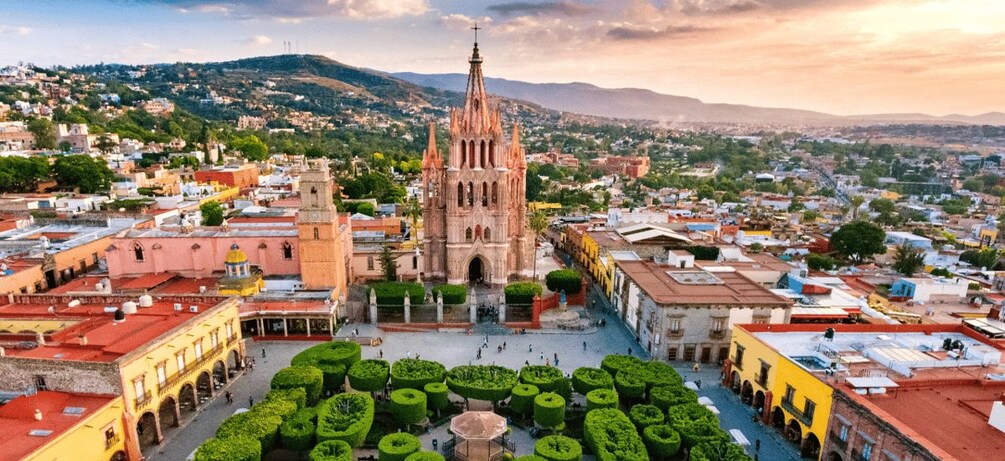 Una catedral con una aguja alta, rodeada por una plaza con setos recortados y un espacio público con gente. 1