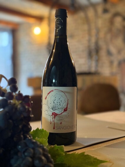 A bottle of wine with a label featuring a grape cluster and a flower, placed on a table with grapes and leaves around it. 5