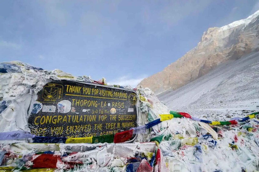 Un letrero con un mensaje de felicitación por el trekking en Manang, rodeado de nieve y coloridas banderas de oración. 1