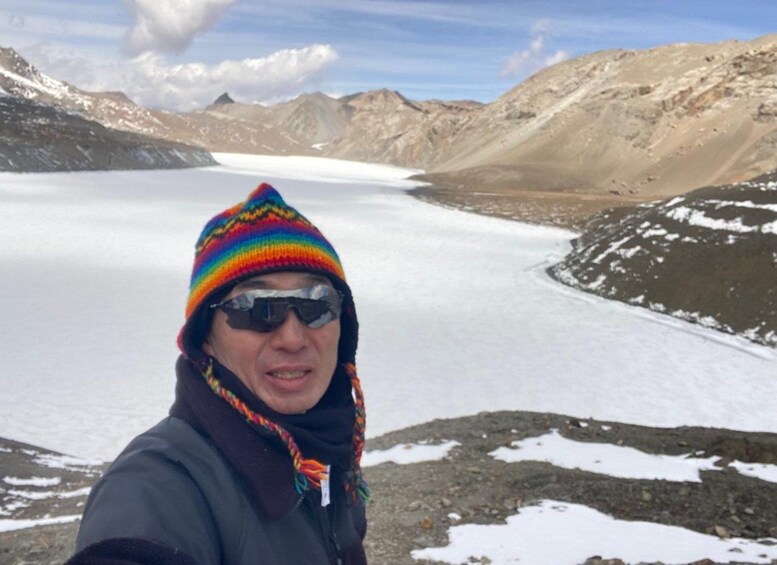 Una persona con un gorro de lana colorido y una chaqueta oscura de pie frente a un glaciar, con montañas al fondo. 5