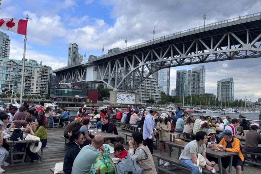 A crowded outdoor dining area with people sitting at tables, a bridge in the background, and a city skyline. 1