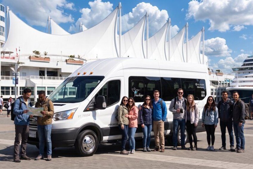 A group of people standing in front of a white van with a sail-like structure on top, parked on a paved area with buildings in the background. 5